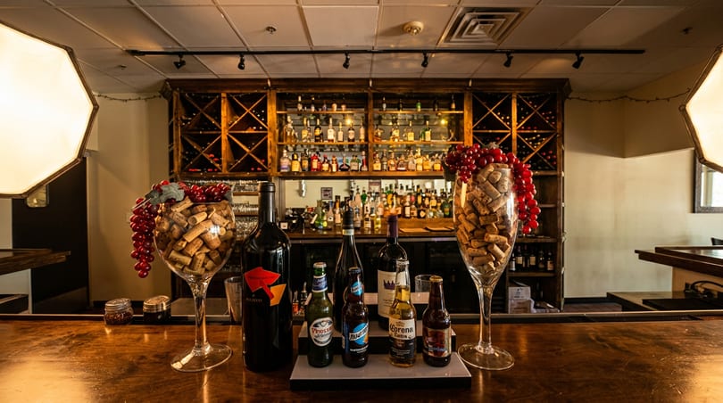 A bar counter with wine bottles, beer bottles, and large glasses filled with corks and grapes.