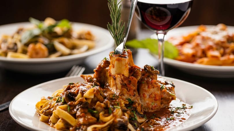 Osso buco with tagliatelle pasta, red wine, and other pasta dishes on a dark table.