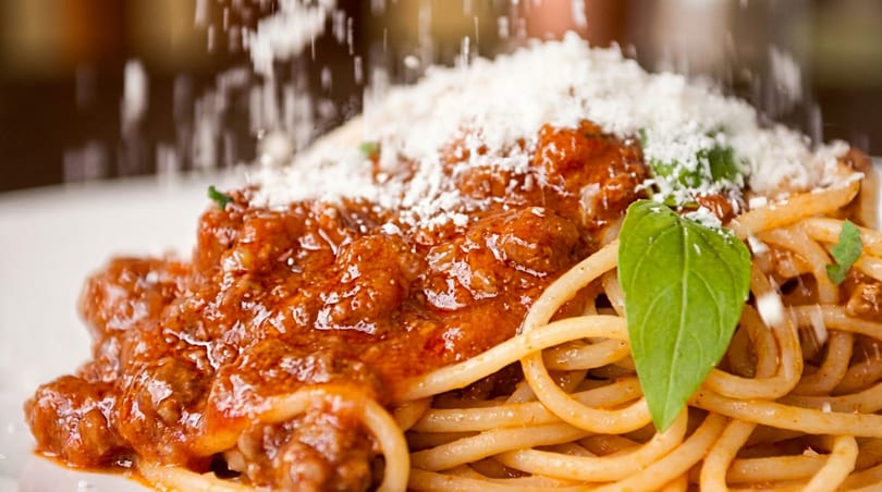 Close-up of spaghetti bolognese with fresh basil, being sprinkled with grated Parmesan cheese.