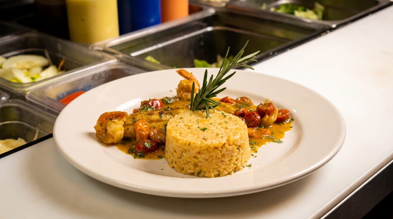 Plated seafood dish with rice, shrimp, fish, cherry tomatoes, and rosemary garnish in a restaurant kitchen.