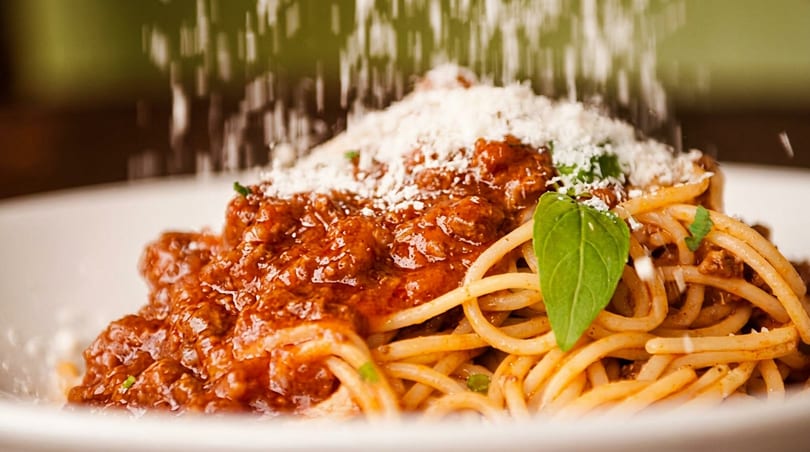 Parmesan cheese being sprinkled over a plate of spaghetti bolognese with a basil leaf garnish.
