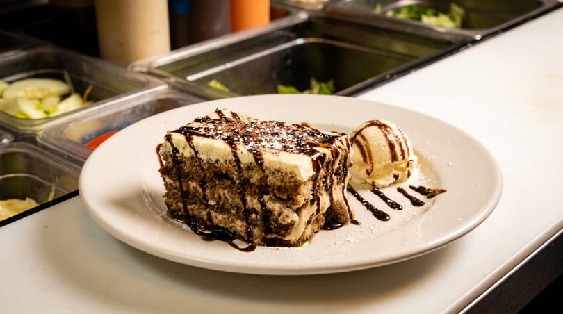A slice of tiramisu with chocolate drizzle and a scoop of vanilla ice cream on a white plate.