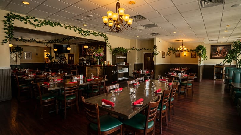 An empty restaurant dining room with tables set, a bar in the background, and ivy decorations.